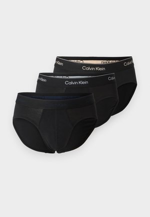 Τρία μαύρα ανδρικά εσώρουχα Calvin Klein με ελαστικές ζώνες μέσης με λογότυπο, στοιβαγμένα σε λευκό φόντο.
