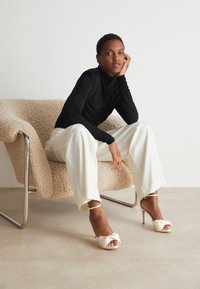 Black turtleneck, white wide-leg trousers, and satin peep-toe heels with bow detail. The model sits on a textured beige armchair.