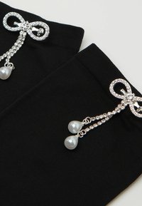 Sorte leggings med en dekorativ sølvsløjfe-brosche med rhinsten og to perle-lignende dråber, der er fastgjort med delikate kæder.