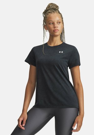 Jonge vrouw met gevlochten haar, gekleed in een zwart Under Armour T-shirt en grijze legging, staand met één hand op de heup tegen een witte achtergrond.