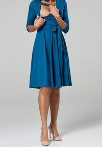 Tyrkysové zavinovací šaty s tříčtvrtečními rukávy, se zavázaným pasem a rozšířenou sukní. V kombinaci s nahými vysokými podpatky. Hladký textilní povrch.
