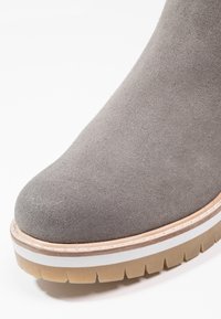 Gros plan sur le bout d'une botte en daim gris avec une semelle superposée comportant des bandes de roulement en caoutchouc beige, blanc et marron avec des rainures profondes pour la traction.