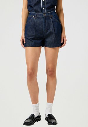 Shorts de mezclilla en azul oscuro con cintura alta, con pliegues frontales, dos bolsillos laterales y remaches de cobre. Combinados con mocasines negros y calcetines blancos.