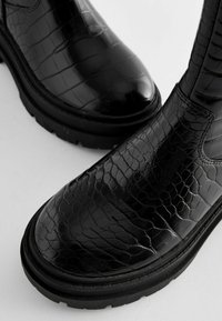 Schwarze Knöchelschuhe mit Reptilstruktur, glänzender Oberfläche und Gummisohle. Sie haben eine glatte Form und elastische seitliche Einsätze für ein einfaches Anziehen.
