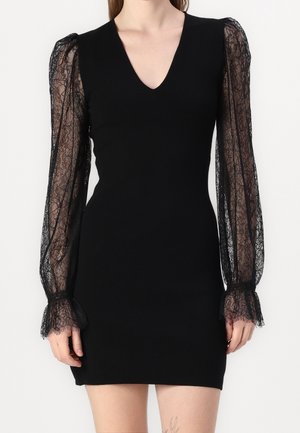 Czarna dopasowana sukienka z dekoltem w serek i przezroczystymi koronkowymi długimi rękawami z falbanianymi mankietami, noszona przez osobę stojącą na jednolitym tle.