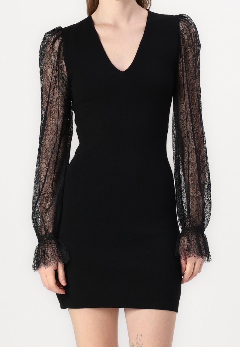 Robe noire ajustée avec col en V et manches longues en dentelle transparente avec des poignets à volants, portée par une personne debout sur un fond uni.