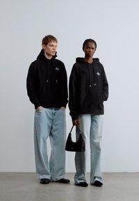 Dvaja modely v čiernych mikinách s kapucňou a logom, doplnených svetlomodrými džínsami s širokými nohavicami. Jeden z nich drží malú čiernu kabelku.