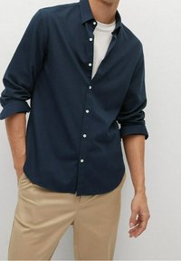 Uomo che indossa una camicia button-up blu navy su una maglietta bianca con maniche arrotolate e pantaloni beige, in piedi con una mano in tasca.