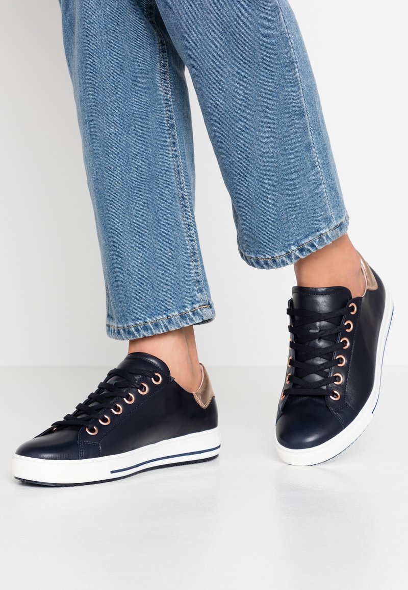 Personne portant un jean bleu et des baskets basses noires avec des semelles blanches et des œillets en or rose, debout sur une surface blanche.