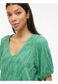 Blusa verde con una texture a V e maniche corte a sbuffo. Il tessuto presenta un motivo increspato, creando un effetto visivo dinamico.