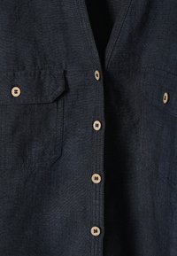 Gros plan sur le devant d'une chemise en tissu foncé avec des boutons en bois clair et deux poches poitrine boutonnées avec rabats.