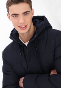 Tengerészkék steppelt kabát kapucnival, cipzáras záródással, rugalmas mandzsettákkal és sima felülettel. A modell karjait összefonta egy fehér háttér előtt.