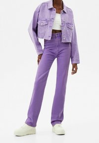 Przycięta fioletowa kurtka dżinsowa z dwoma kieszeniami na piersi oraz pasującymi fioletowymi dżinsami. Model nosi biały top na ramiączkach i jasne kremowe sneakersy.