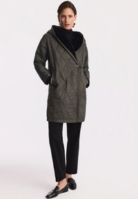 Pikowane oliwkowe płaszcz z dużym kapturem i czarnym futrem w środku, z bocznym paskiem i dwiema kieszeniami, w zestawieniu z czarnymi spodniami i loafersami.