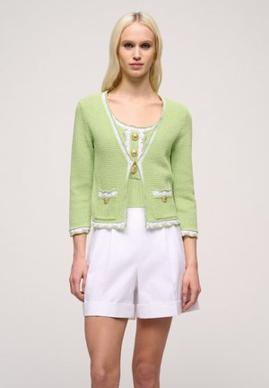 Cardigan tricotat verde deschis cu detalii albe, cu mâneci de trei sferturi și două buzunare; asortat cu un top corespunzător și pantaloni scurți albi.