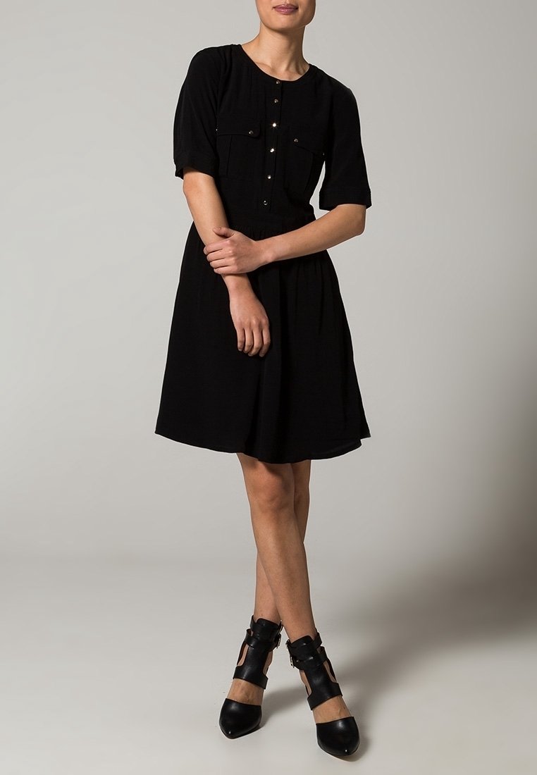 Femme debout, jambes croisées, portant une robe noire mi-longue à boutons et des talons noirs à bride découpée sur fond gris.