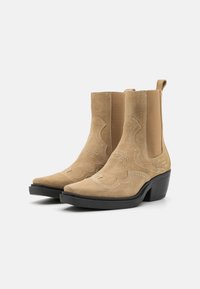 Botines de ante color beige con puntera afilada, que presentan costuras decorativas y paneles laterales elásticos, montados sobre un tacón alto negro.