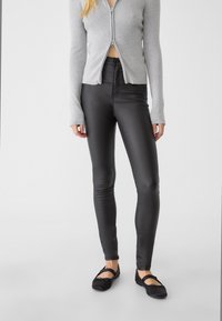Zwarte, aansluitende neplederen broeken met een gladde textuur en een hoge taille. Gecombineerd met een lichtgrijze geribbelde cardigan met rits.