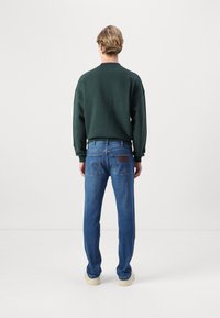 Sudadera verde oversized, jeans azules con un parche de cuero, combinados con zapatillas de color claro. Vista trasera, fondo blanco liso.