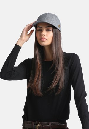 Jeune femme aux longs cheveux raides porte une casquette grise et un pull noir, touchant la visière de la casquette avec sa main droite.