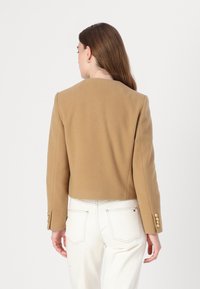 Brun cropped jakke i blødt stof med rund halsudskæring og guldknapsdetaljer på ærmerne, parret med hvide bukser.