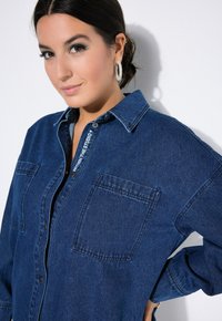 Camicia di jeans di un blu profondo, con colletto button-down, due tasche sul petto e un dettaglio del logo lungo la patta. Tessuto morbido e vestibilità comoda.
