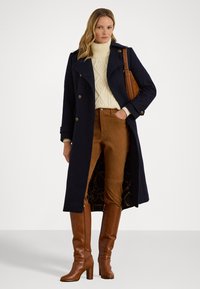 Abrigo de lana azul marino con botones de doble botonadura sobre un suéter de punto trenzado color crema y pantalones de gamuza marrón, combinados con botas marrones hasta la rodilla.