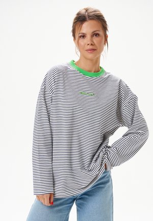 Jeune femme aux cheveux bruns portant une chemise rayée à manches longues avec un col vert et un jean bleu, debout avec la main sur la hanche sur fond blanc.