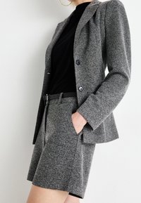 Blazer grigio testurizzato con bottoni neri abbinato a pantaloni corti su misura, con una tasca e una cintura nera. Sotto una maglietta nera.