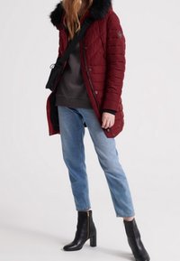 Quiltet rød jakke med en faux fur hætte, parret med en grå sweatshirt og blå jeans. Sorte ankelstøvler med guldaccent fuldender looket.