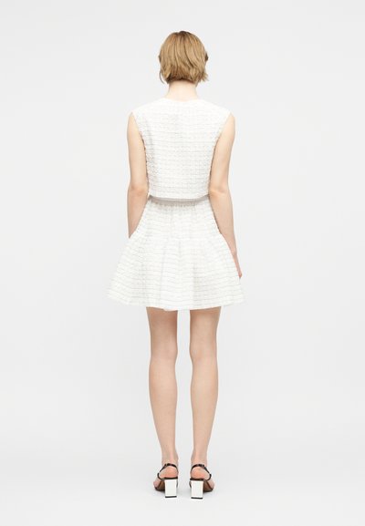 Άτομο με κοντά ξανθά μαλλιά, που φοράει λευκό αμάνικο φόρεμα με υφή και μαύρα πέδιλα με χοντρό τακούνι και λουράκια, στέκεται με την πλάτη προς τα εμπρός.