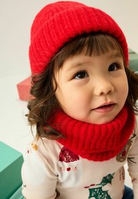 Un jeune enfant aux cheveux ondulés porte un bonnet et une écharpe en tricot rouge vif, levant le regard avec une expression joyeuse, entouré de boîtes cadeaux colorées.