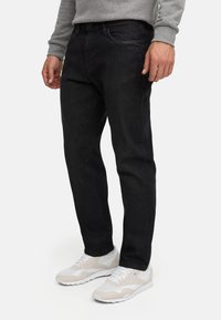 Mann iført svarte jeans og hvite joggesko står på hvit bakgrunn med hendene avslappet ned langs sidene.