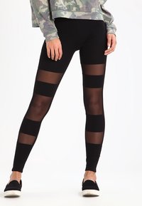 Leggings noirs avec des panneaux rayés transparents. Texture lisse avec un design ajusté. Associés à un sweat-shirt camouflage et à des chaussures noires à enfiler.