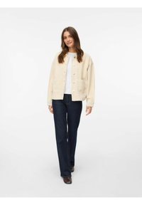 Beige jacka med knappar, två framfickor, bärs över en vit topp och mörkblåjeans. Slät textur, avslappnad passform och långa ärmar.
