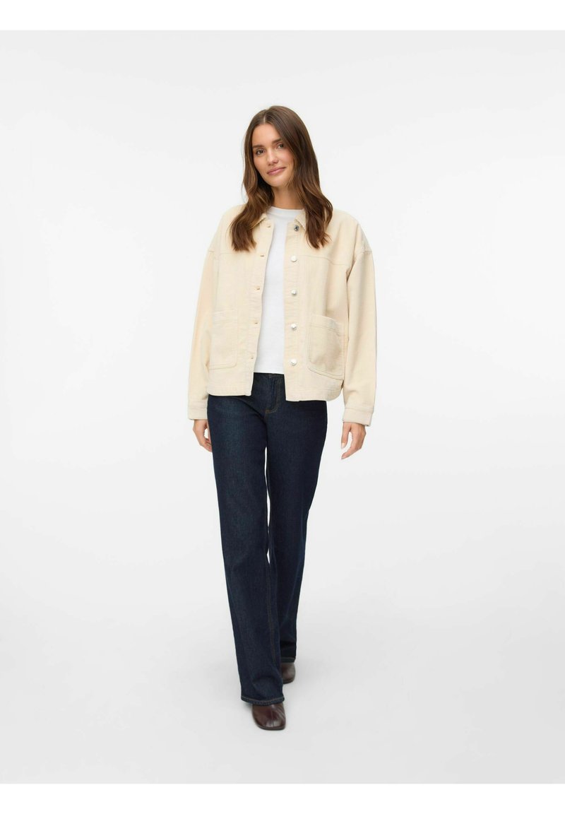 Beige jacka med knappar, två framfickor, bärs över en vit topp och mörkblåjeans. Slät textur, avslappnad passform och långa ärmar.