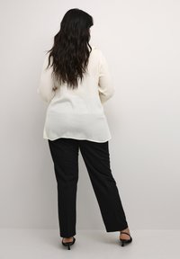 Langærmet beige bluse i tekstureret stof, parret med sorte skræddersyede bukser. Blusen har en buet kant og en afslappet pasform.