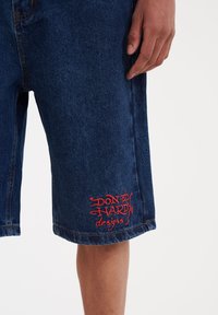 Denim shorts in dark blue with a relaxed fit. Features red embroidered text "DON ED HARDY designs" on the left leg, slight fraying at the hem.