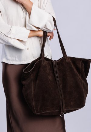 Person trägt eine weiße Bluse und einen braunen Rock und hält eine große dunkelbraune Wildledertasche mit zwei Schulterriemen.
