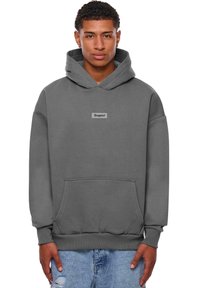 Graue Kapuzenjacke mit einer Tasche vorne, verstellbarem Kordelzug an der Kapuze und einem rechteckigen Logo-Patch in der Mitte. Hergestellt aus weichem, strukturiertem Stoff.