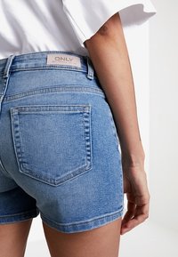Pantalones cortos de mezclilla azul con un corte clásico, dos bolsillos traseros y una etiqueta con "ONLY" en la cintura. Textura suave, lavado medio.