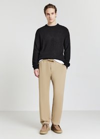 Junger Mann mit nach hinten gebundenem Haar, der einen schwarzen Strickpullover, beige Hosen und braune Wildlederschuhe trägt, vor einem schlichten weißen Hintergrund steht.