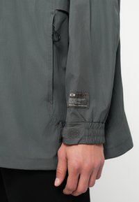 Κοντινή λήψη μανικιού γκρι πανωφοριού με ελαστική μανσέτα και λουράκι Velcro, που δείχνει μία ετικέτα της μάρκας Oakley σε άσπρο και μαύρο πάνω από τον καρπό.