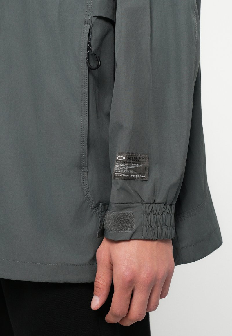 Close-up van een grijze jas mouw met elastische manchet en klittenbandsluiting, met een zwart-witte Oakley merklabel boven de pols.