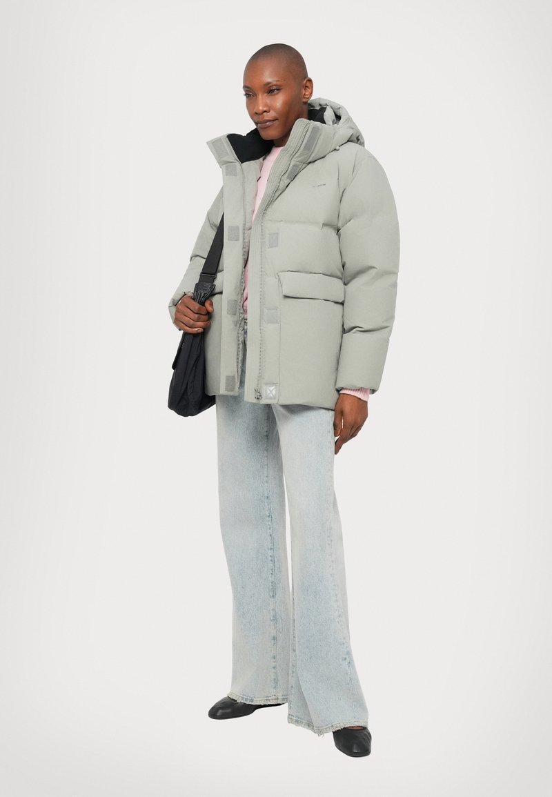 Veste grise rembourrée avec capuche, plusieurs poches frontales et accents gris. Associée à un jean évasé bleu clair et des chaussures noires. Sac noir.