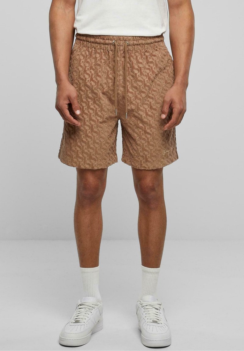 Homme portant un short marron texturé avec un cordon de serrage, des chaussettes blanches montantes et des baskets blanches, debout devant un fond gris uni.