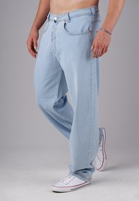 Bare-chested person wearing light blue loose-fit jeans and white low-top sneakers, standing against a plain gray background.