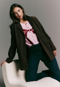 Brown, suede-style jacket over a pink sweater with a red graphic design. Dark blue jeans. Model posed on a white sculptural chair.