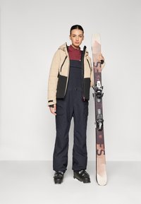 Beige-schwarze Daunenjacke über einem dunkelroten Oberteil, kombiniert mit dunklen Latzhosen. Hält ein Snowboard mit schwarzen Bindungen und schwarzen Skischuhen.