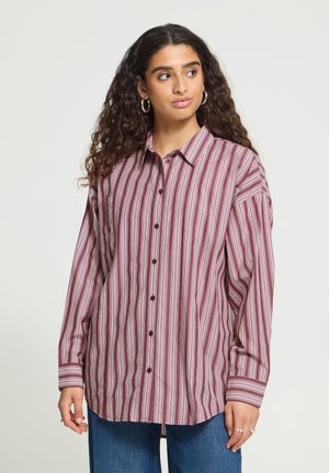 Giovane donna con lunghi capelli ricci indossa una camicia oversize a righe verticali rosse e bianche con bottoni e jeans blu, guardando verso destra.